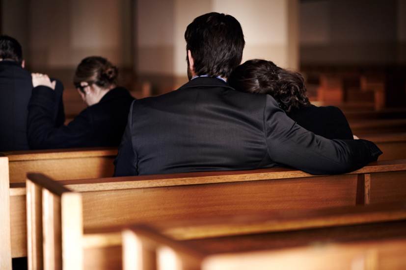 Mourners attend a funeral. A couple sits in a wooden pew with the woman leaning on the man's shoulder in a somber, supportive moment.
